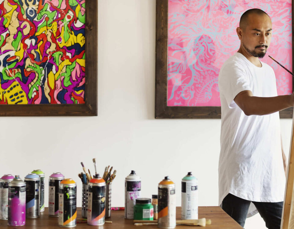 Artista plástico concentrado pintando em um ateliê, cercado por latas de tinta spray, pincéis e quadros coloridos ao fundo. Cena representa criatividade, expressão artística e ambiente de produção visual contemporânea.